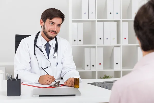 Doctor smiling to his patient and writing in clippad. They sit in doctors office. Concept of doctor patient relationship
