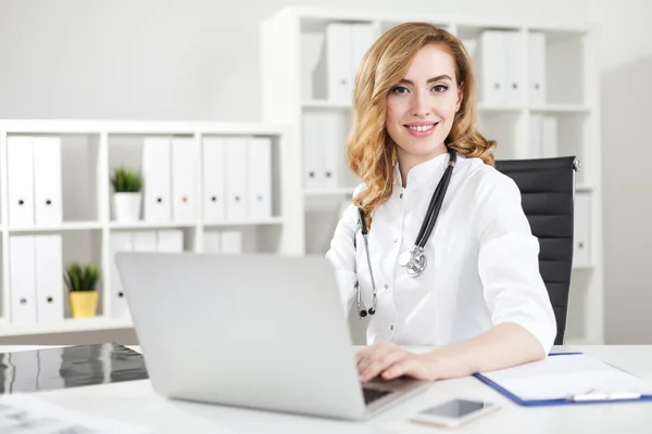 Cheerful woman doctor typing on her laptop keyboard sitting in white uniform in her office and smiling. Concept of good mood