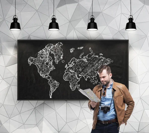 Handsome tourist is examining the map in front of chalkboard with the world map sketch