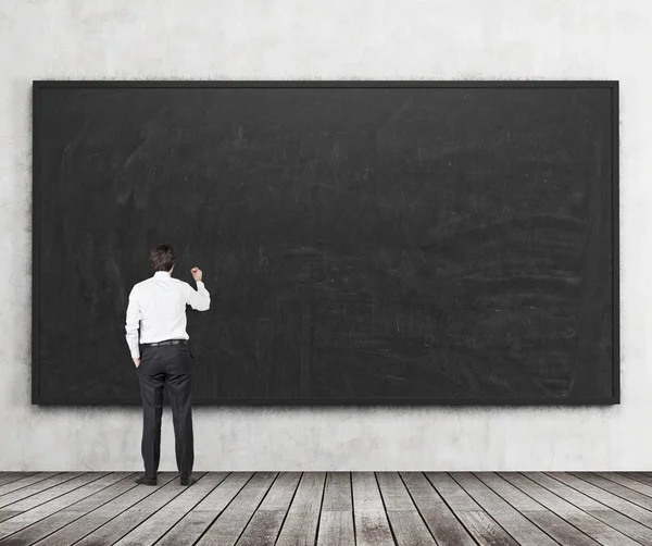 Rear view of the man who is going to write something on the black chalkboard. Wooden floor and concrete wall. A concept of the beginning of new academic year. A class room.