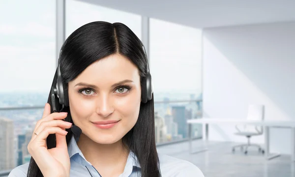Front view of the smiling brunette support phone operator with headset. Panoramic office workplace on the background. New York city view.