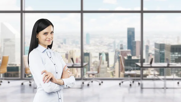 A side view of a beautiful smiling lady with crossed hands. The brunette woman is dressed in formal clothes. Modern workplaces in a fancy panoramic office in New York.