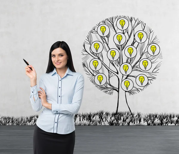A smiling beautiful brunette is pointing out something by her hand. A sketch of a tree with light bulbs is drawn on the concrete wall. Light bulbs as a concept of new business ideas.