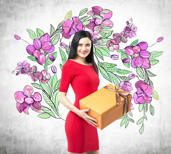 A smiling brunette in a red dress holds an orange gift box. The sketch of purple flowers is drawn on the concrete wall.