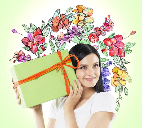 A curious brunette woman tries to guess what is inside the green gift box. The sketch of colourful flowers is drawn on light green wall.