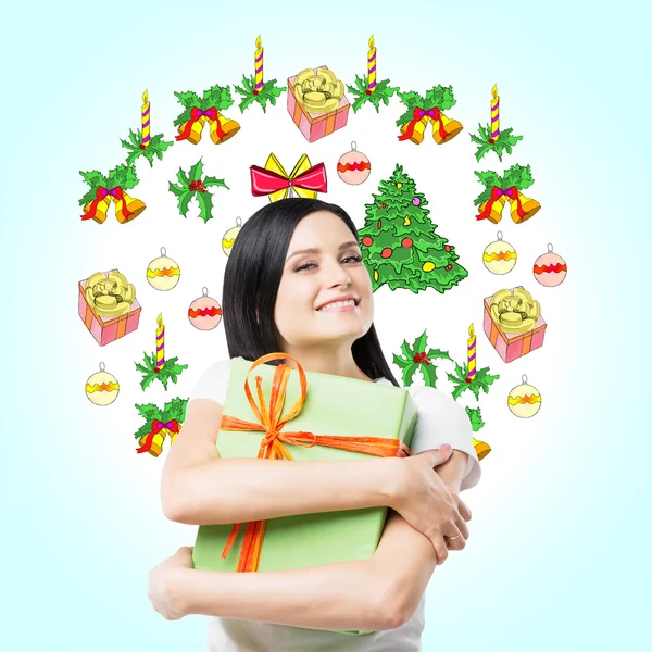 A happy brunette woman is hugging a green gift box. Christmas-tree decorations are drawn on the light blue background. A concept of Christmas and New Year's Eve.