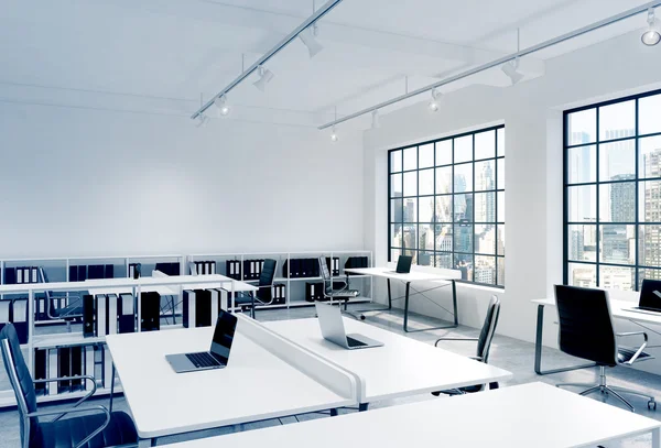 Workplaces in a bright modern loft open space office. Tables equipped with laptops; corporate documents\' shelves. New York in the panoramic windows. 3D rendering.