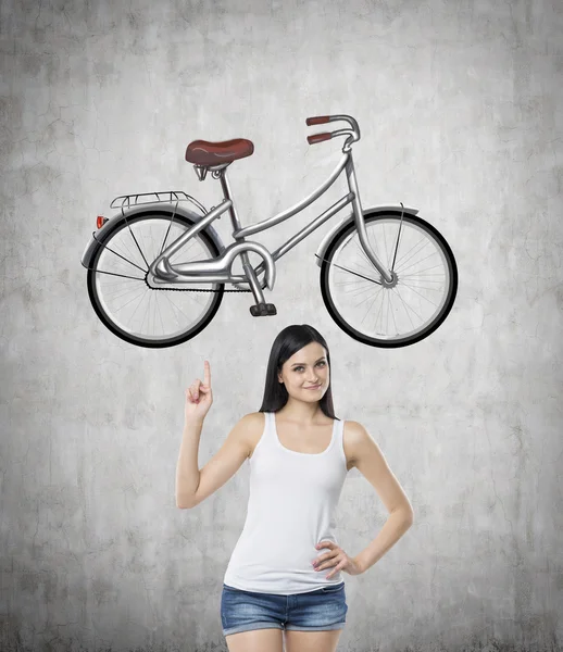A brunette girl in a white tank top and denim shorts points out the bicycle by her finger. A sketch of bicycle is drawn on the concrete wall.