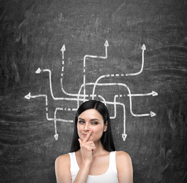 A portrait of a young artful brunette lady who is trying to find out the solution of some problem. The lady is in a white tank top. Arrow with different directions are drawn on the black chalkboard on