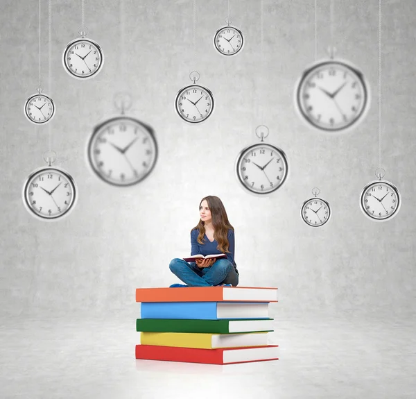 Young woman sitting on pile of books with hanging clocks around