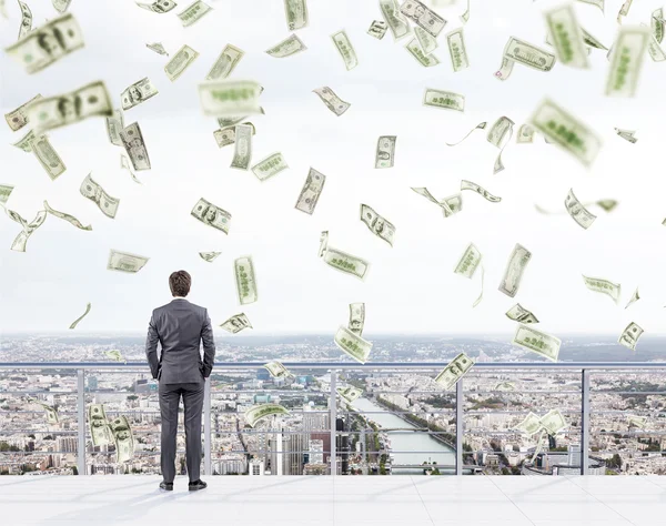 Young businessman with hands in pockets standing on the roof of a business centre and looking at New York. Money falling from above.