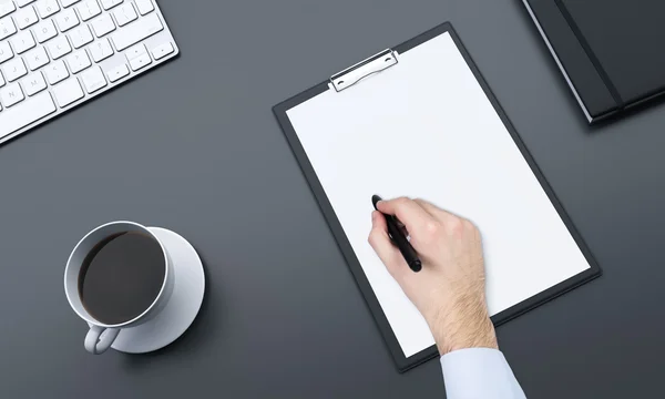 Fragment of a workplace with blank notepad and a cup of black coffee to the left, datebook to the right, hand making notes with a pen.