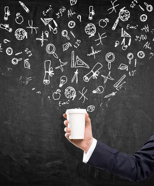 A hand in a black suit holding a paper cup. Black background with different scientific symbols drawn over the cup.