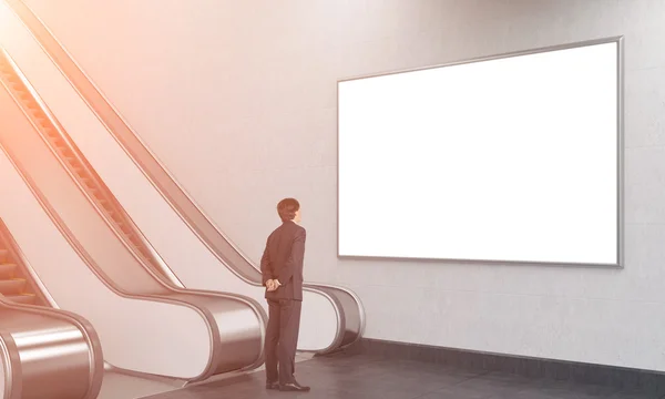 Young businessman with his hands at the back looking at a blank poster over an escalator in the underground.