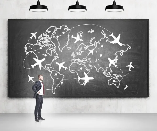 A man standing with hands on hips by a blackboard with planes flying around the globe drawn on it.