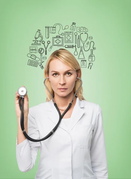 A young female doctor in a white smock holding a phonendoscope and standing in front of the green wall, many medical icons drawn on it.