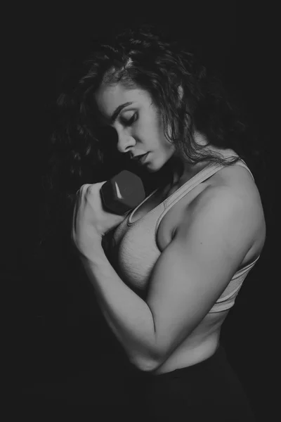 Black and white photo of beautiful athlete on a dark background. Women's fitness. Working with dumbbells. Photos for sporting magazines, posters and websites.
