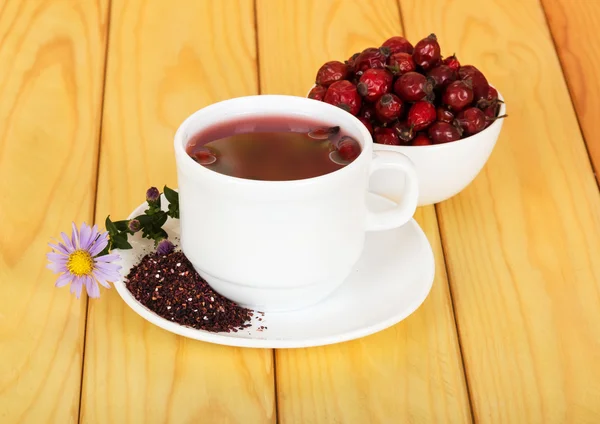 Bowl  dog rose berries, cup tea on  background light wood.