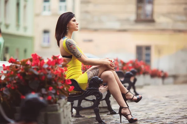 A beautiful woman in a yellow dress, tattoos, sits on the bench,