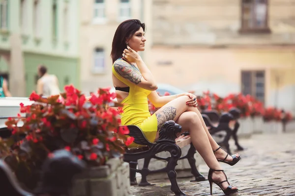 A beautiful woman in a yellow dress, tattoos, sits on the bench,