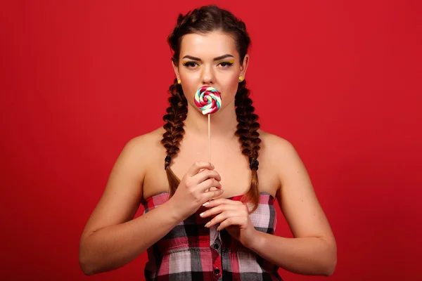 Beauty fashion model girl eating colorful lollipop. Surprised funny young woman with braids hairstyle, nails and makeup beige, isolated on a red background