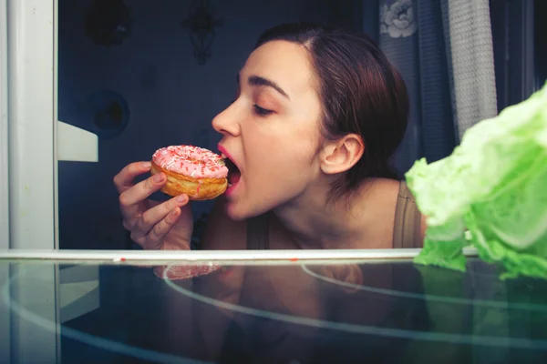 Hungry attractive girl eating donut at night near fridge