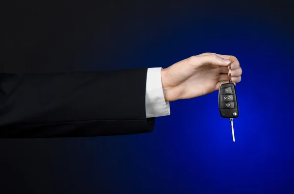 Business and gift theme: car salesman in a black suit holds the keys to a new car on a dark blue background in studio