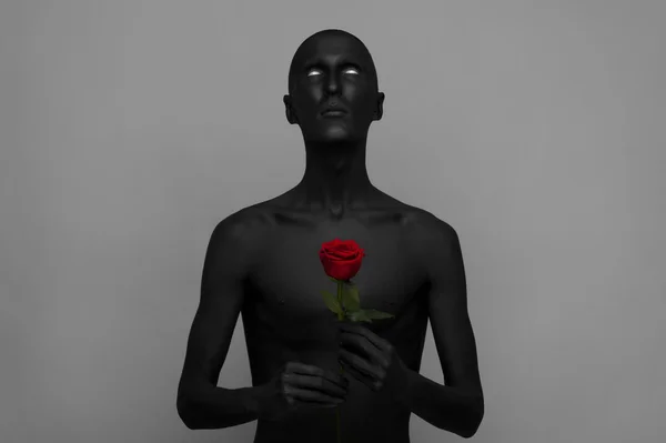 Gothic and Halloween theme: a man with black skin holding a red rose, black death isolated on a gray background in studio