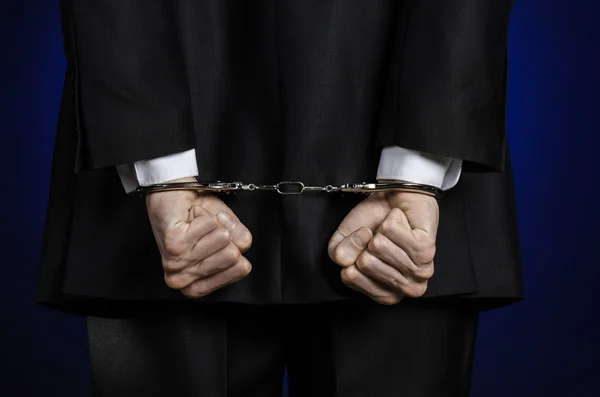 Corruption and bribery theme: businessman in a black suit with handcuffs on his hands on a dark blue background in studio isolated view from the back