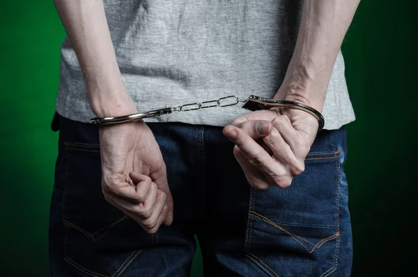 Prison and convicted topic: man with handcuffs on his hands in a gray T-shirt and blue jeans on a dark green background in the studio, put handcuffs on the drug dealer, the view from the back
