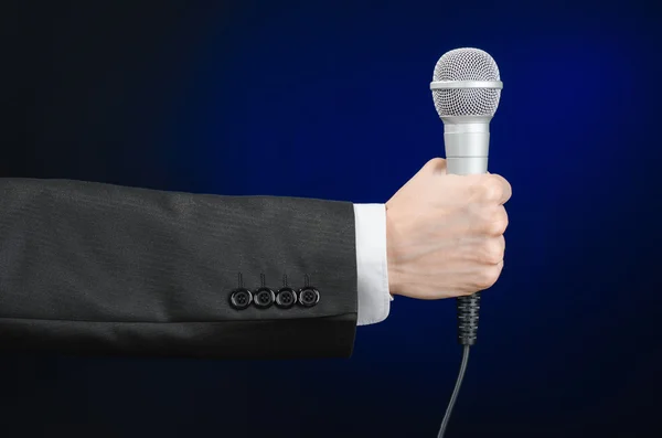 Business and speech topic: Man in black suit holding a microphone on a gray dark blue isolated background in studio