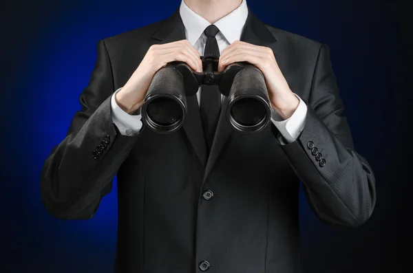 Business and search topic: Man in black suit holding a black binoculars in hand on a dark blue background in studio isolated