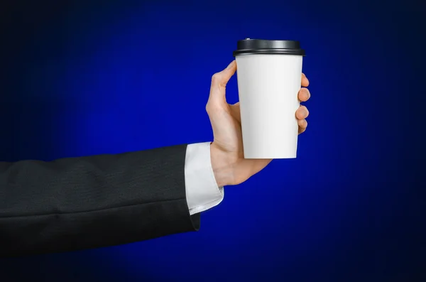 Business lunch and coffee theme: businessman in a black suit holding a white blank paper cup of coffee with a brown plastic cap on a dark blue background isolated in the studio, advertising coffee