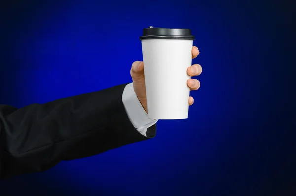 Business lunch and coffee theme: businessman in a black suit holding a white blank paper cup of coffee with a brown plastic cap on a dark blue background isolated in the studio, advertising coffee