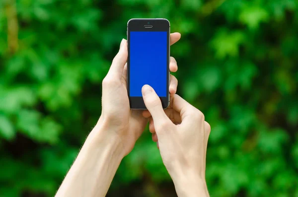 Human hand holding a modern mobile phone with a blue screen on a background of green grass first-person view