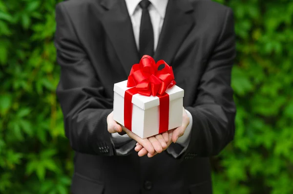 Gift and business theme: a man in a black suit holding a gift in a white box with a red ribbon on a background of green grass
