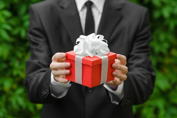 Gift and business theme: a man in a black suit holding a gift in a red box with a white ribbon on a background of green grass