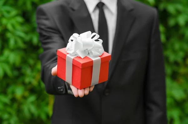 Gift and business theme: a man in a black suit holding a gift in a red box with a white ribbon on a background of green grass