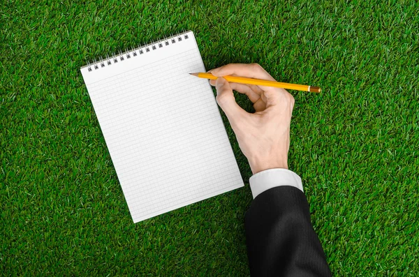 Day of knowledge and business topic: the hand of man in a black suit holding a notebook and pencil top view on a background of green grass