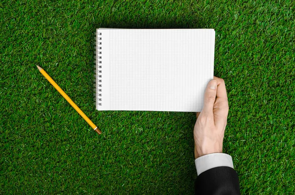 Day of knowledge and business topic: the hand of man in a black suit holding a notebook and pencil top view on a background of green grass