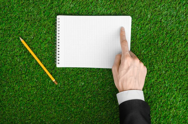 Day of knowledge and business topic: the hand of man in a black suit holding a notebook and pencil top view on a background of green grass