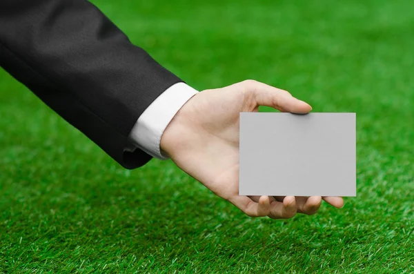 Discounts and business topic: hand in a black suit holding a gray blank card on green grass background