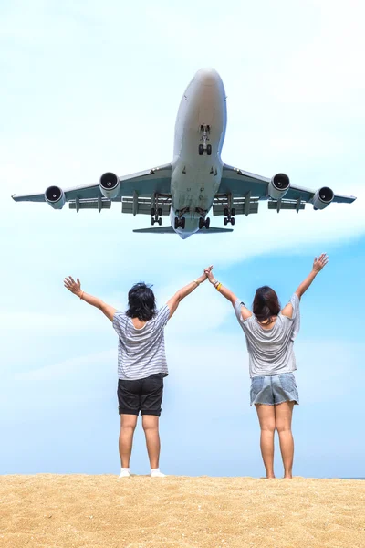 Asian womans raise hands on the beach with airplane landing over head.