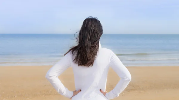 Woman watching the sea, her hands on her hips, seen from the back
