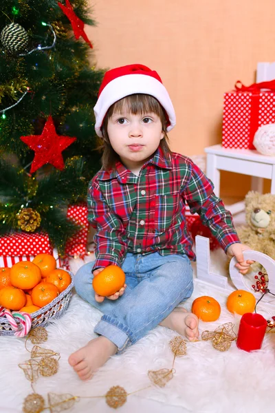 Little boy in Santa hat with tangerine sits near Christmas tree