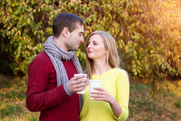 Love, relationship, season, friendship and people concept - happy man and woman enjoying golden autumn fall season with paper coffee cups in the park. Young couple drinking hot cofee or tea.