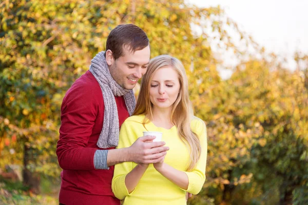 Love, relationship, season, friendship and people concept - happy man and woman enjoying golden autumn fall season with paper coffee cups in the park. Young couple drinking hot cofee or tea.