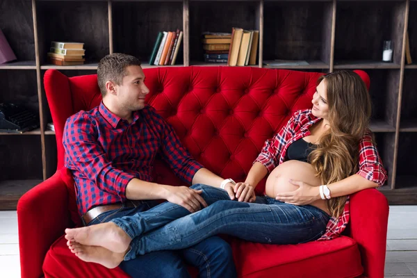 Beautiful pregnant couple relaxing on sofa at home together. Happy family, man and woman expecting a child.