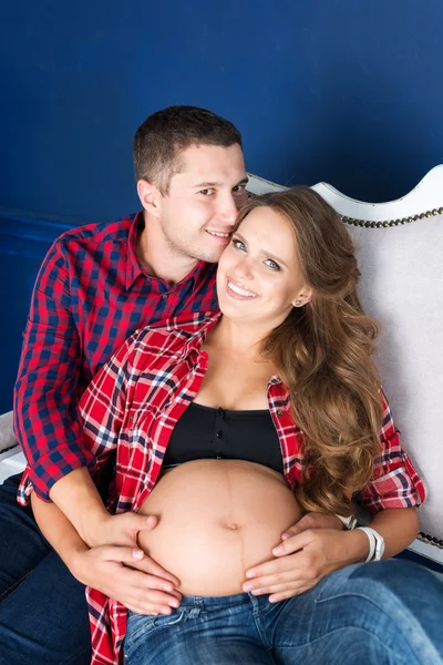 Beautiful pregnant couple relaxing on sofa at home together. Happy family, man and woman expecting a child.