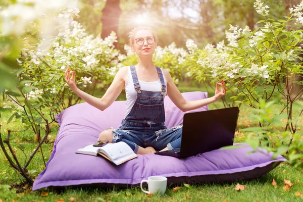 Young business woman doing yoga outside office building sitting in lotus position in the park with her laptop and cup of tea or coffee. Freelance, nature, relaxation concept.